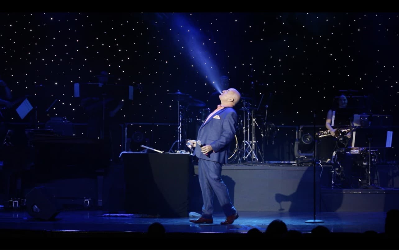 Scott Record on a cruise ship stage under a follow-spot, head back mid-gag, the full band behind him